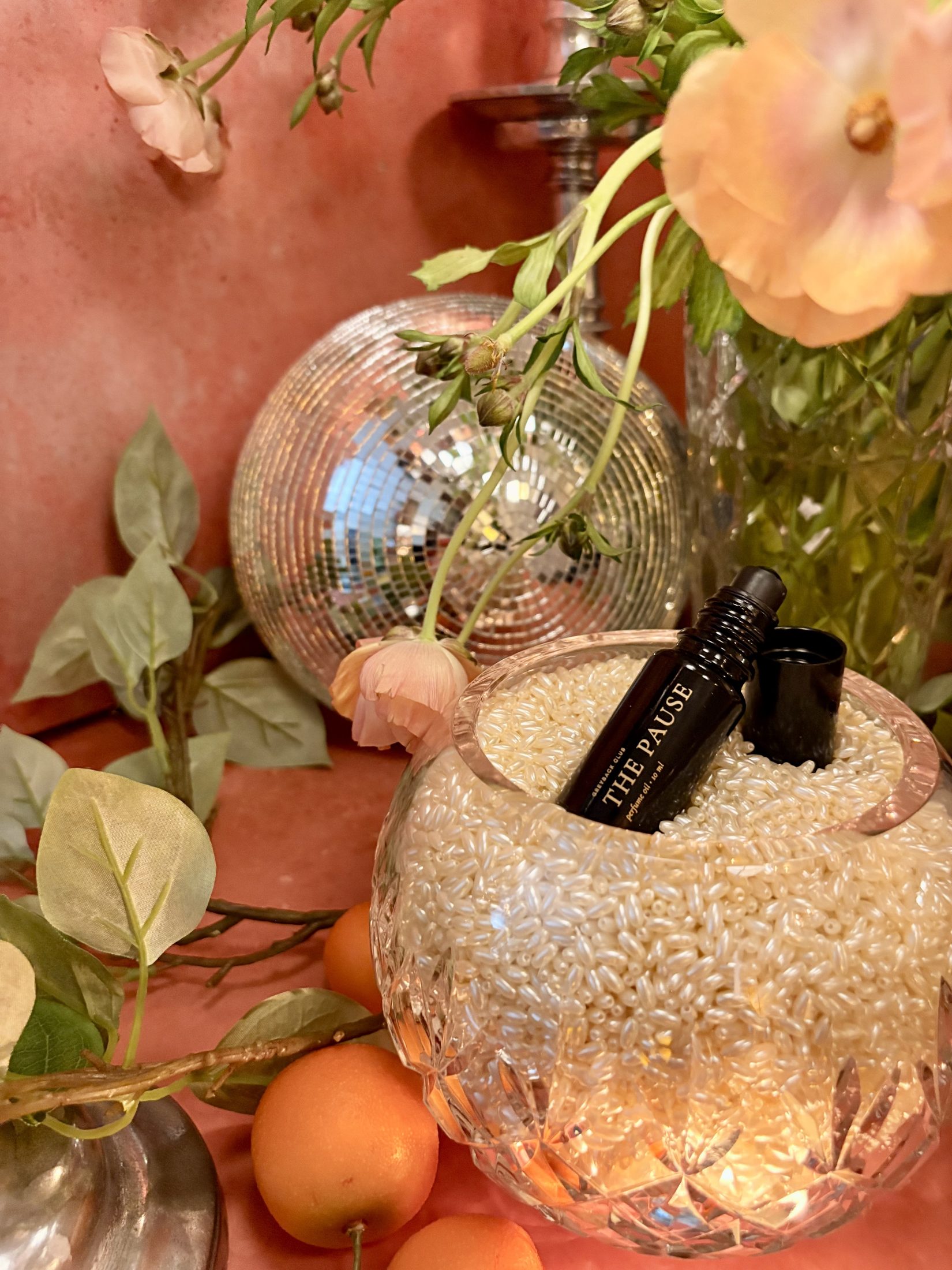 The Pause oil resting in a crystal bowl of pearls, with peach flowers, a disco ball and kumquats